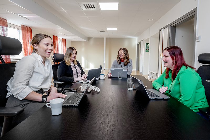 Fyra personer sitteri vid ett bord i kontorsmiljö med datorer och skrattar.